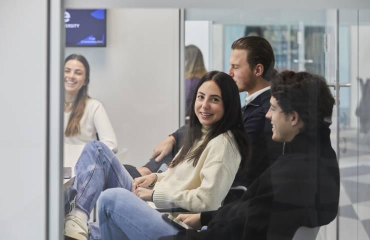 Four young adults are seen sitting and chatting casually inside a modern office environment.