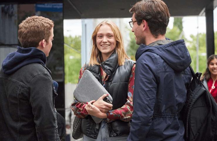 Three young adults are having a conversation at a bus stop, with the female in the center holding a laptop and smiling.