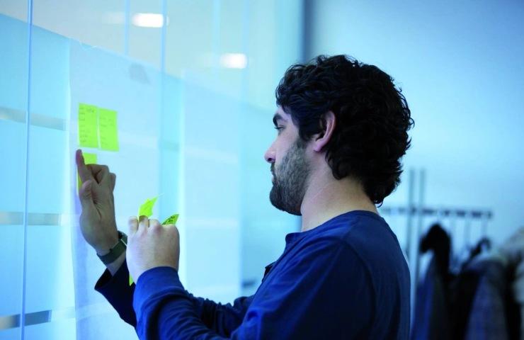 A man is attaching sticky notes to a glass wall in an office setting.