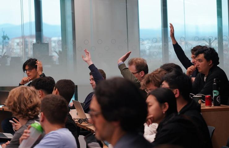 A group of people in a conference room, some of whom are raising their hands.