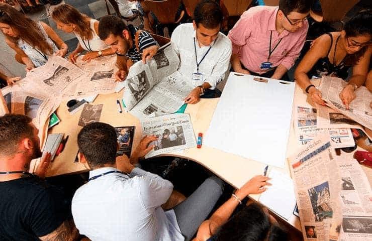 A group of people sitting at a table, engaged in reading and discussing over multiple newspapers spread out before them.