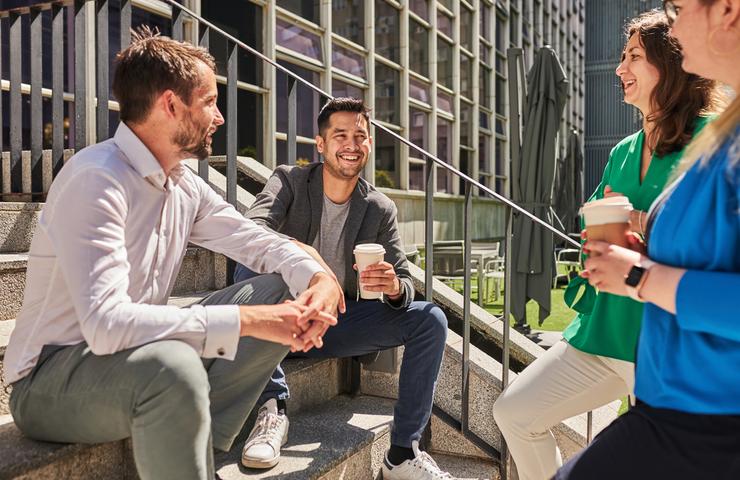 Three people are seated and chatting happily on steps outdoors, with coffee cups in hand, in a sunny urban setting.