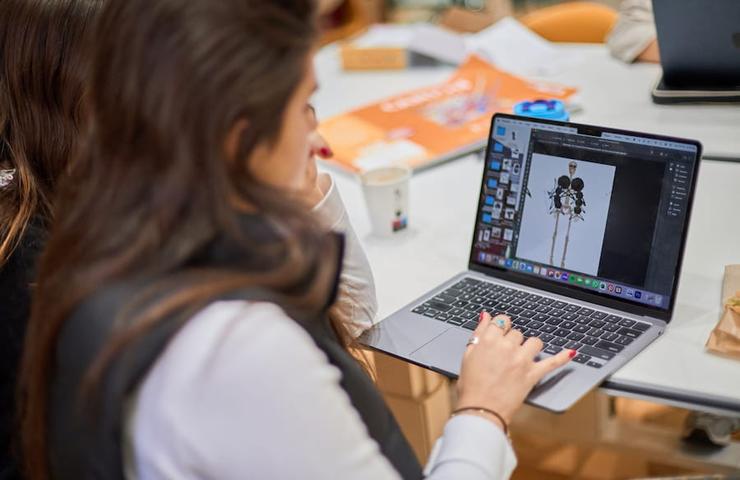 A person working on a laptop with a design software open, surrounded by other items on a table.