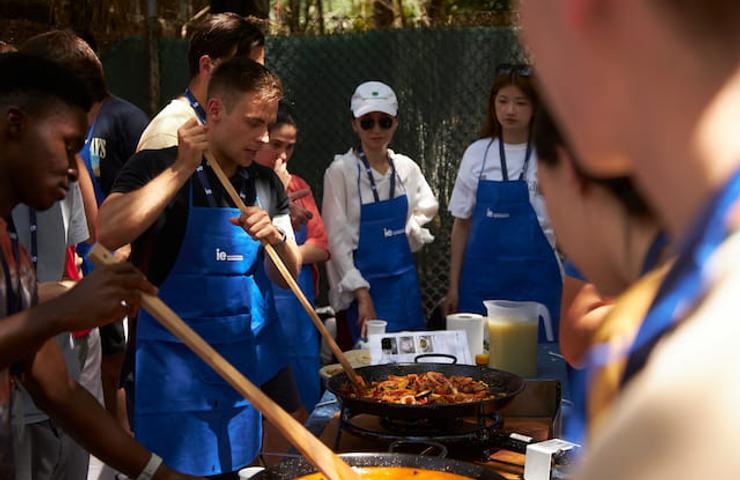 People in blue aprons are cooking and stirring a large paella outdoors.