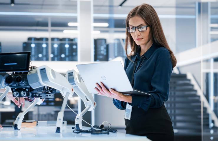 A woman is working on a laptop in a modern tech environment with robotic equipment.