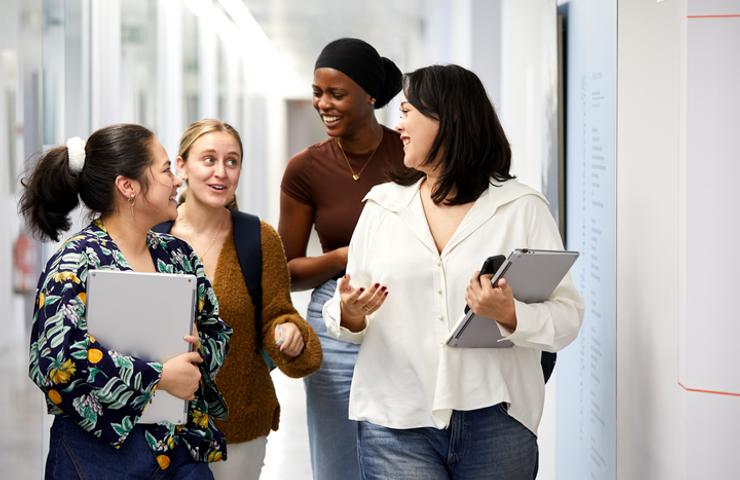 Four women, each holding laptops or notebooks, are engaged in conversation in a bright office corridor.