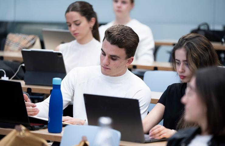 A group of students engaged in a classroom setting using laptops and taking notes.