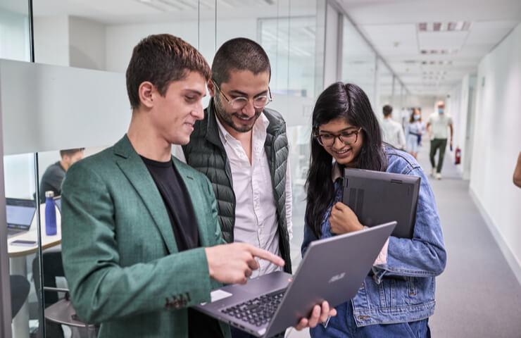 Three professionals, two men and one woman, are discussing over a laptop in a modern office corridor.