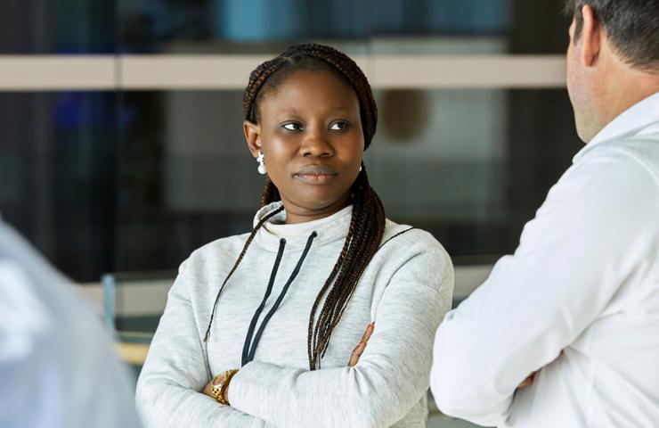 A woman with braided hair stands with her arms crossed, looking thoughtfully towards a man in a business setting.