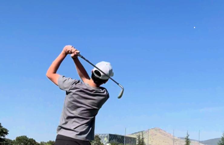 A person in a grey shirt and white cap is swinging a golf club on a sunny day with clear blue skies.