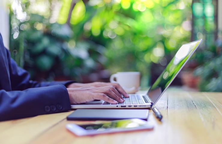 A person working on a laptop at a wooden table outdoors with a blurred green background.