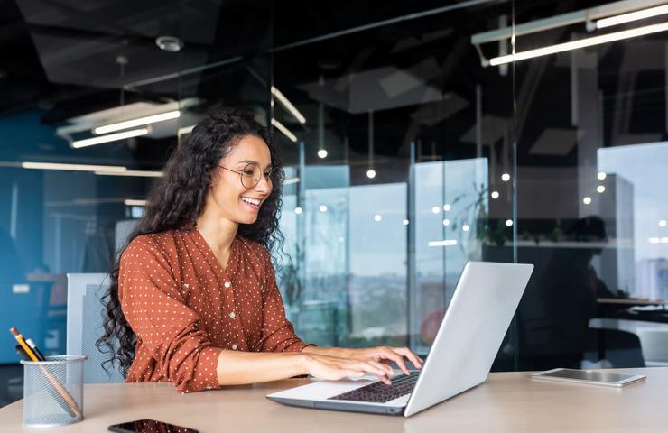 A woman in a polka dot shirt smiling and working on a laptop in a modern office environment.