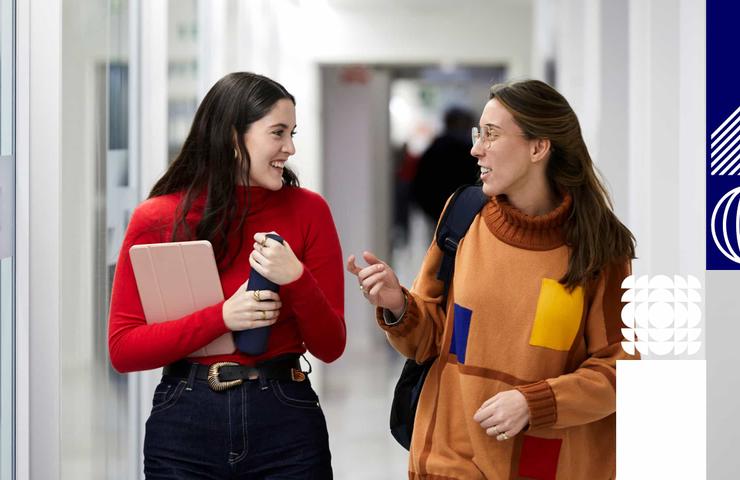 Two young women happily conversing while walking through a school corridor, one holding a notebook and the other a cup.