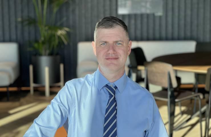 A man in a blue shirt and striped tie smiling at the camera with a modern office background.