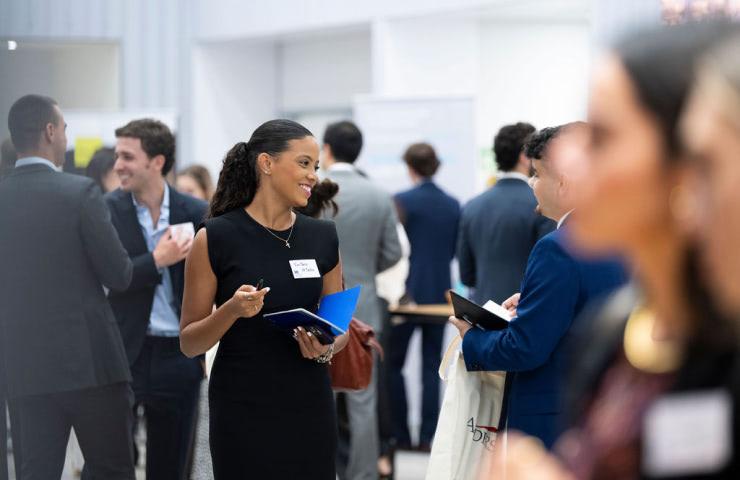 A woman with a name tag smiles and chats at a networking event while holding a notebook.