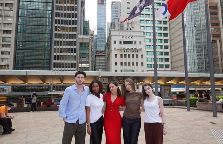 A group of five young adults standing together in an urban setting with tall buildings in the background.