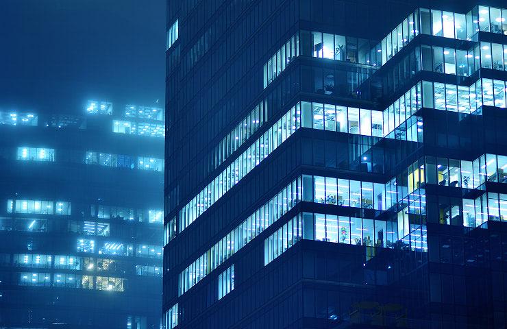 A night scene of modern buildings illuminated by blue lights in a foggy atmosphere.