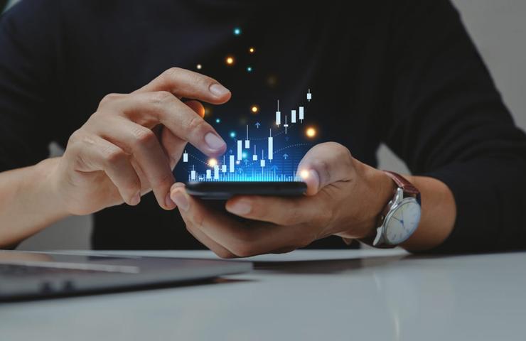 A person analyzing virtual financial data on a smartphone with futuristic holographic statistics, sitting at a desk with a laptop.