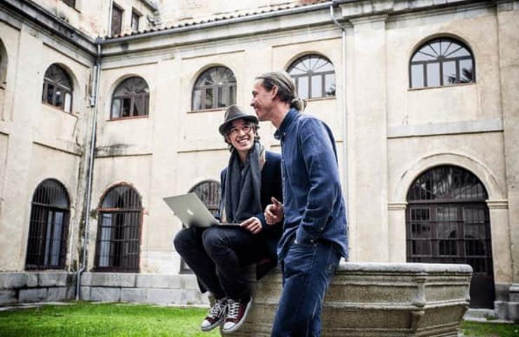 Two men smiling and discussing over a laptop in a garden with historical architecture in the background.