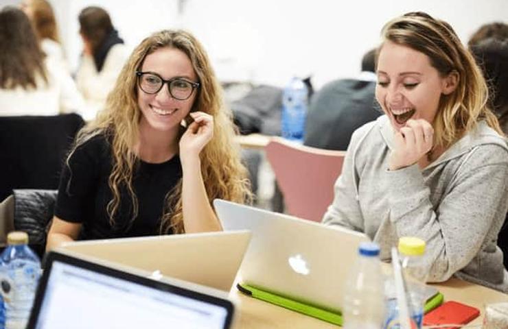 Two women are laughing and looking at a laptop screen in a classroom setting.