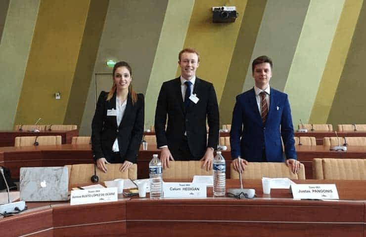 Three young professionals stand behind a desk in a large conference hall, each wearing a name tag and positioned in front of name placards.