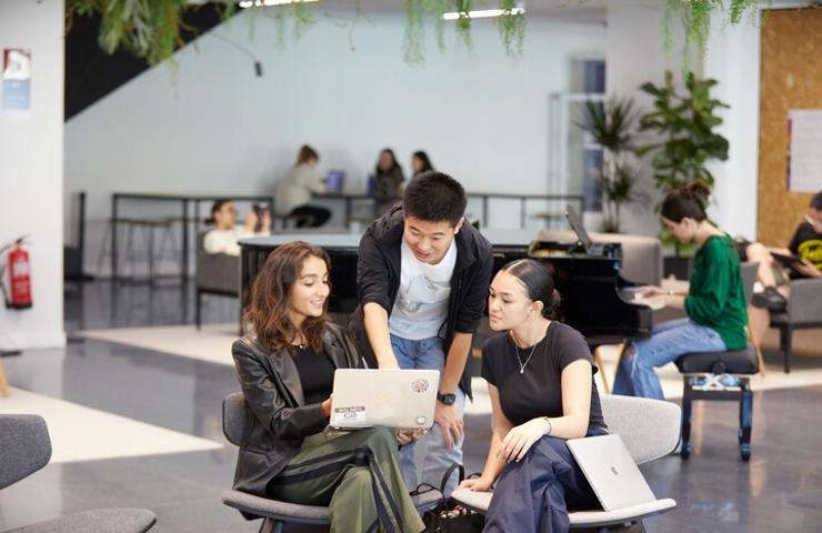 A group of three students collaborating on a laptop in a modern campus setting.