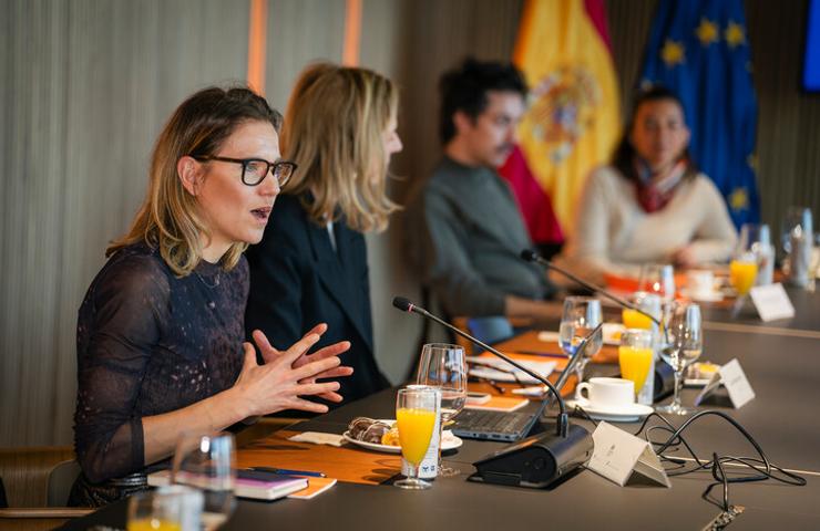 A group of professionals engaging in a discussion at a conference table with beverages and flags in the background.