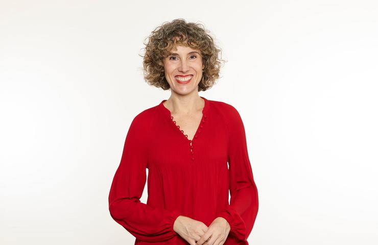 A smiling woman with curly hair wearing a red blouse poses confidently.
