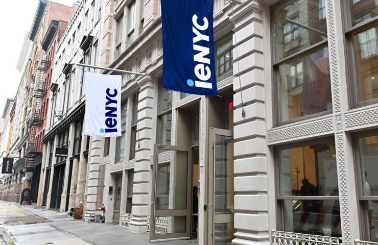 A city street with modern buildings displaying various flags, including one marked 'IENYC'.