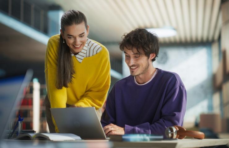 A cheerful collaboration between two young adults working on a laptop in a modern workspace.