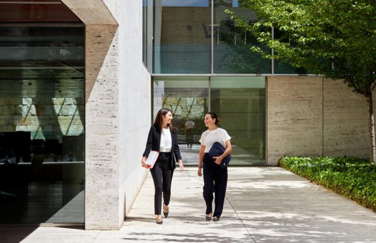 Two women are walking outside a modern building surrounded by greenery.