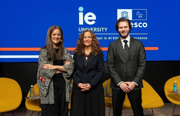 Three people standing on a stage at an event labeled with IE University and UNESCO logos, smiling at the camera.