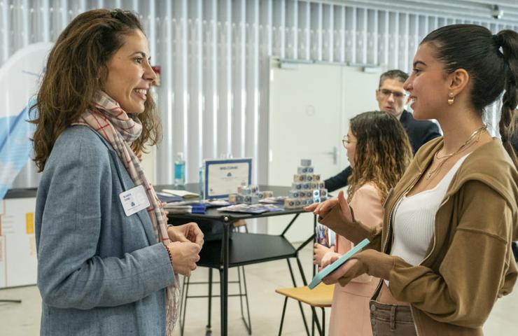 Two women are engaged in a conversation at a business expo, with other attendees and booths in the background.