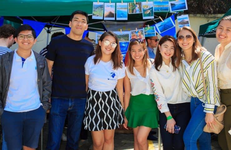 A group of seven diverse people posing happily at an outdoor market or event with several tents and blue-themed decorations in the background.