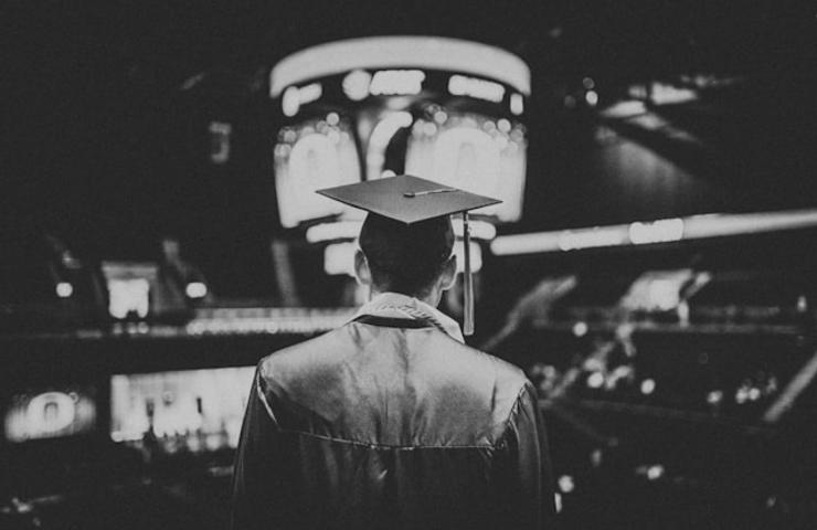 Black and white image of a graduate from behind wearing a cap, facing a blurred stage.