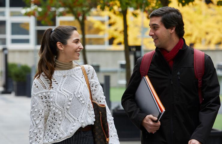Two people are smiling and chatting while walking on a campus with autumn trees in the background.