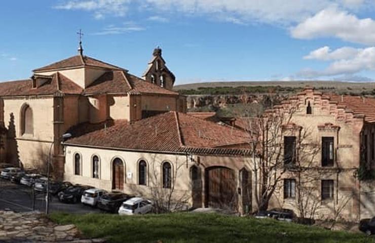 Panoramic view of an ancient monastery complex with terracotta roofs surrounded by sparse vegetation and a clear blue sky.