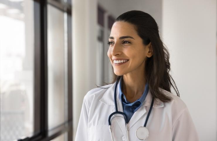 A smiling female doctor wearing a lab coat and stethoscope looks out a window.