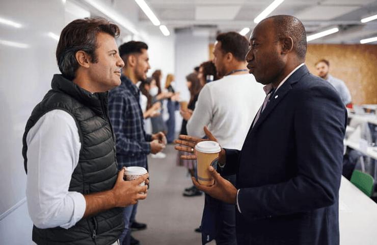 Two men holding coffee cups and talking in an office environment with other people in the background.