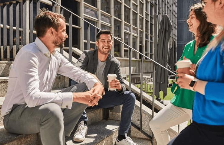 Three colleagues are casually chatting and enjoying coffee on outdoor steps near a modern office building.