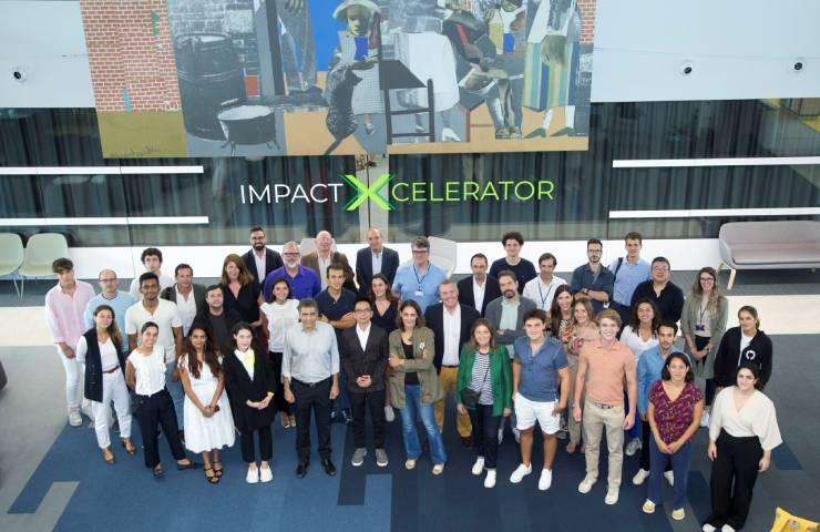 A large group of people posing for a photo in a modern building with a banner reading 'IMPACT XCELERATOR' in the background.