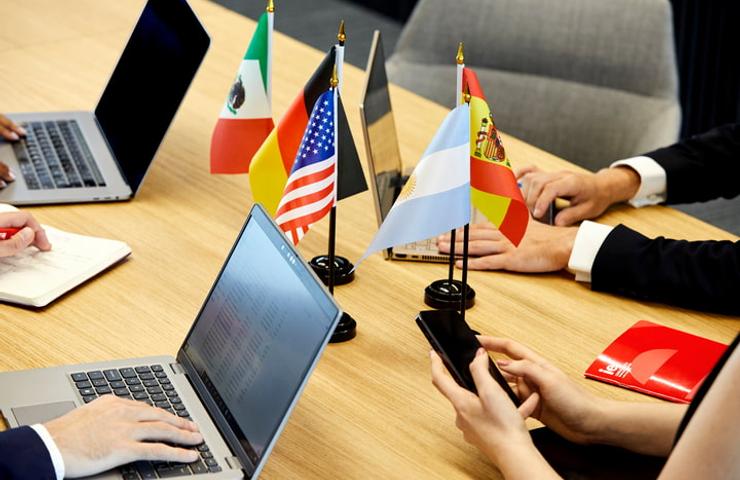 A group of individuals sitting around a table with laptops and various international flags, representing a multinational meeting or conference.