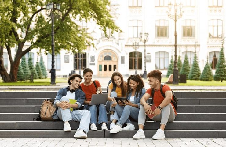 A group of five young people sitting on stone steps, engaging with electronic devices in a park-like setting.
