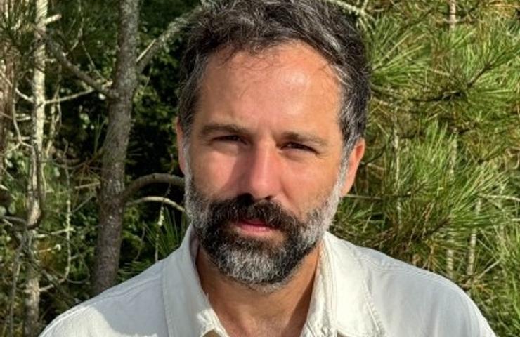 A middle-aged man with a beard and a white shirt standing in front of a wooded area.