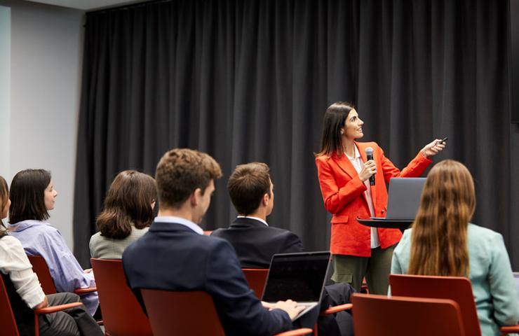 A woman in a red blazer is giving a presentation to a group of seated professionals in a conference room.