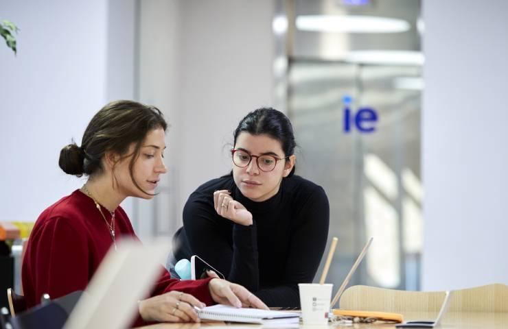 Two women are engaged in discussion while looking at a laptop in a modern office setting.