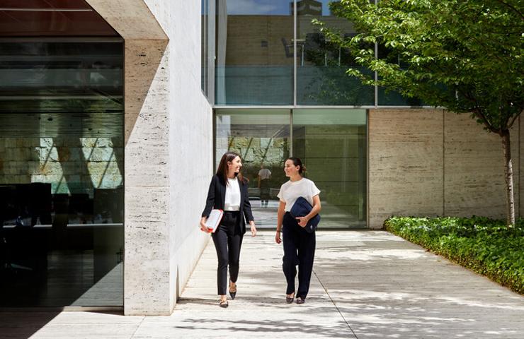 Two women are walking together in a modern architectural setting with greenery.