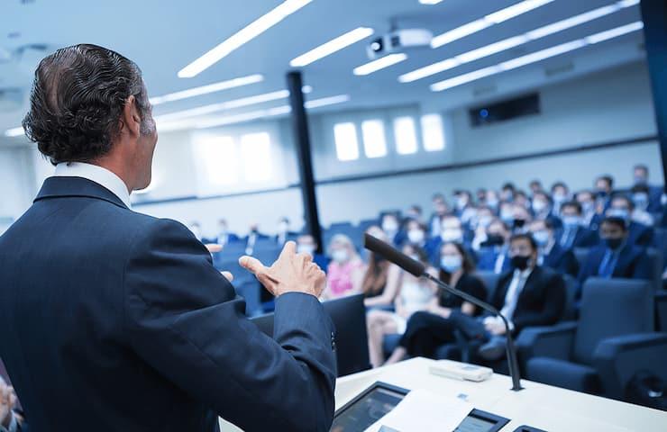 A businessman is giving a presentation in a lecture hall filled with audience members.