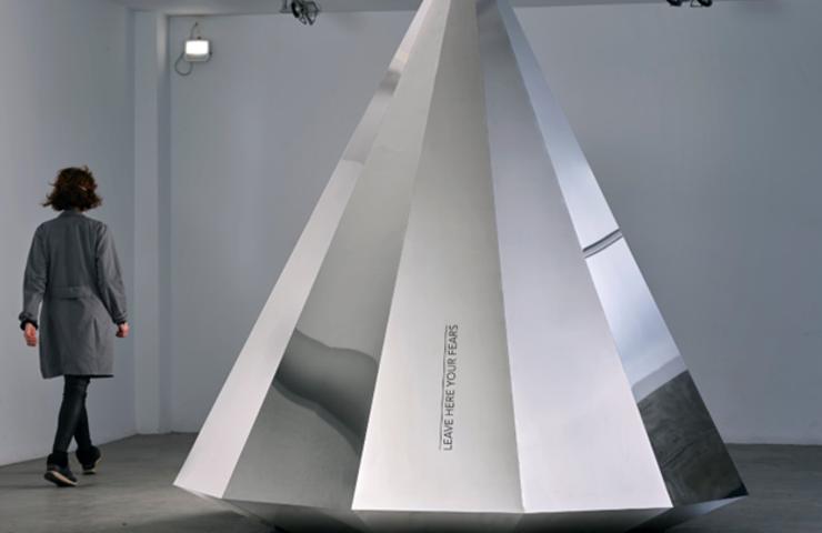 A person observing a large, metallic pyramid structure in a modern art gallery.