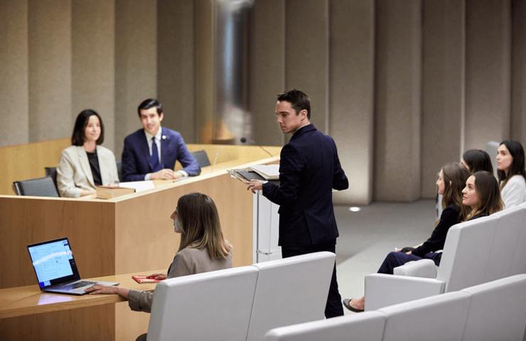 A man speaks at a podium in a modern classroom setting while a group of attentive students and instructors listen.